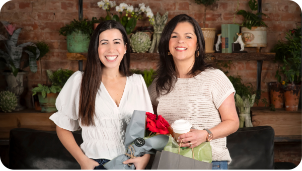 Two ladies stand in a flower shop holding flowers and cups of coffee.